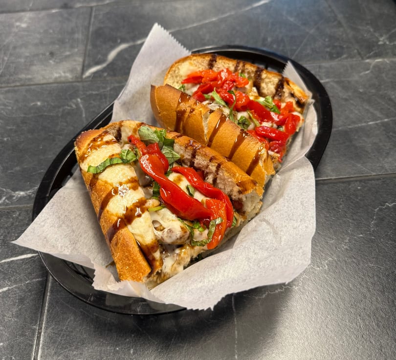 Two halves of a sub sandwich with melted cheese, red peppers, and basil, drizzled with sauce.